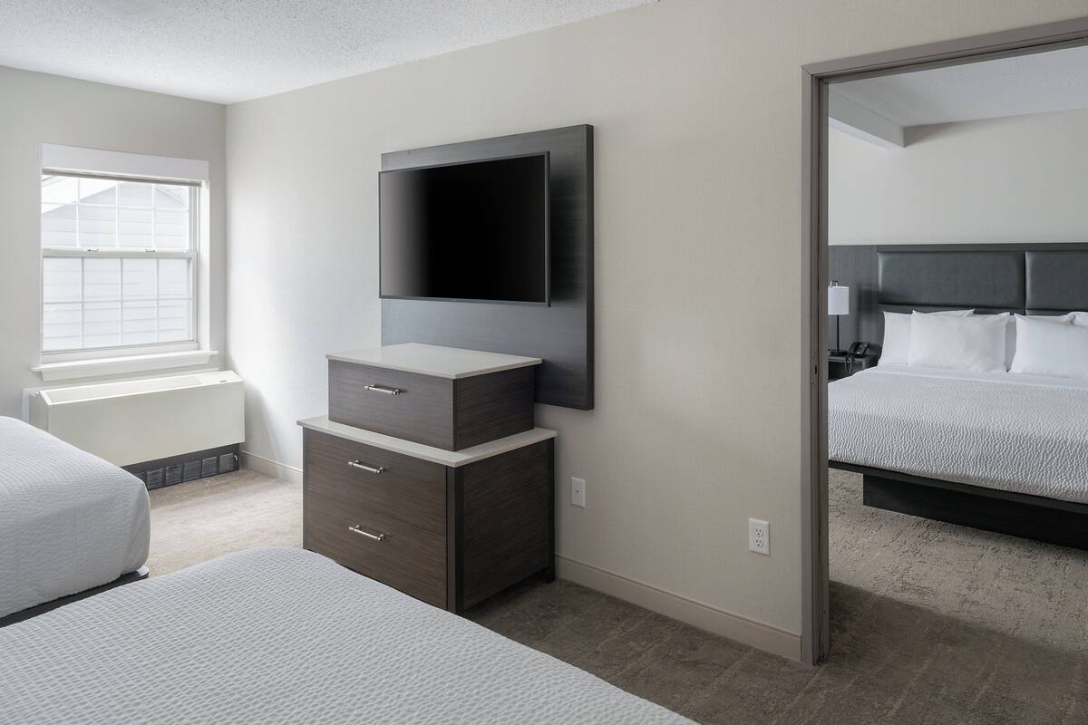 A modern hotel room with a large flat-screen TV mounted on the wall, a bed with white bedding, a bedside table with a lamp, and a window providing natural light.