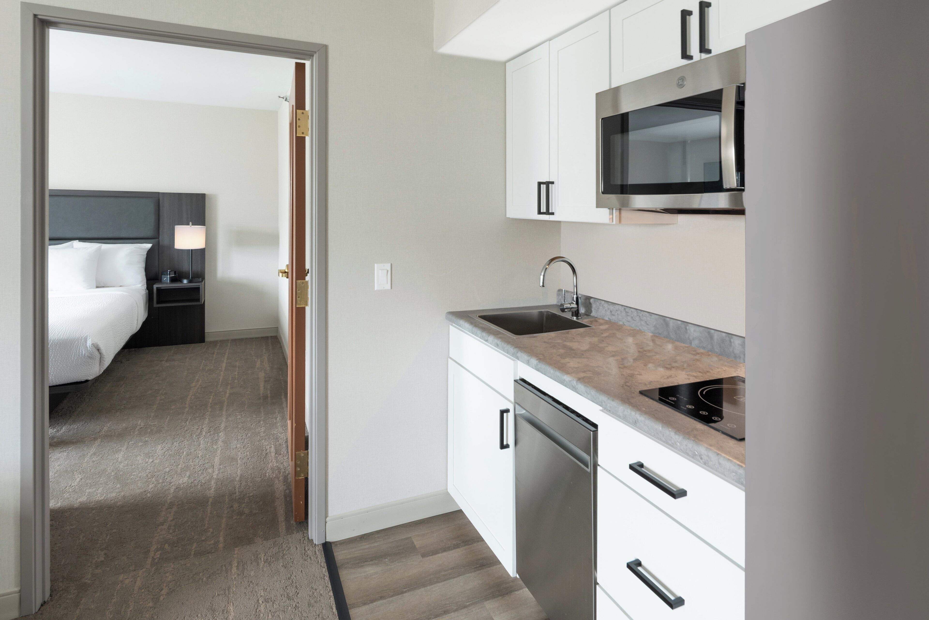 A compact hotel room with a bed, bedside table, lamp, and a kitchenette with a microwave, sink, and dishwasher.