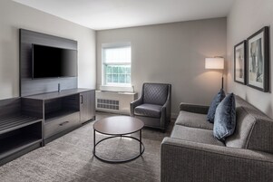 Una sala de estar moderna con un televisor de pantalla plana, un sillón gris, un sofá con cojines, una mesa de centro redonda y una ventana con luz natural.