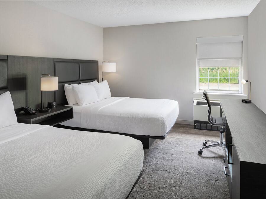 A hotel room with two beds, a desk, a chair, a lamp, and a window with a view of greenery.