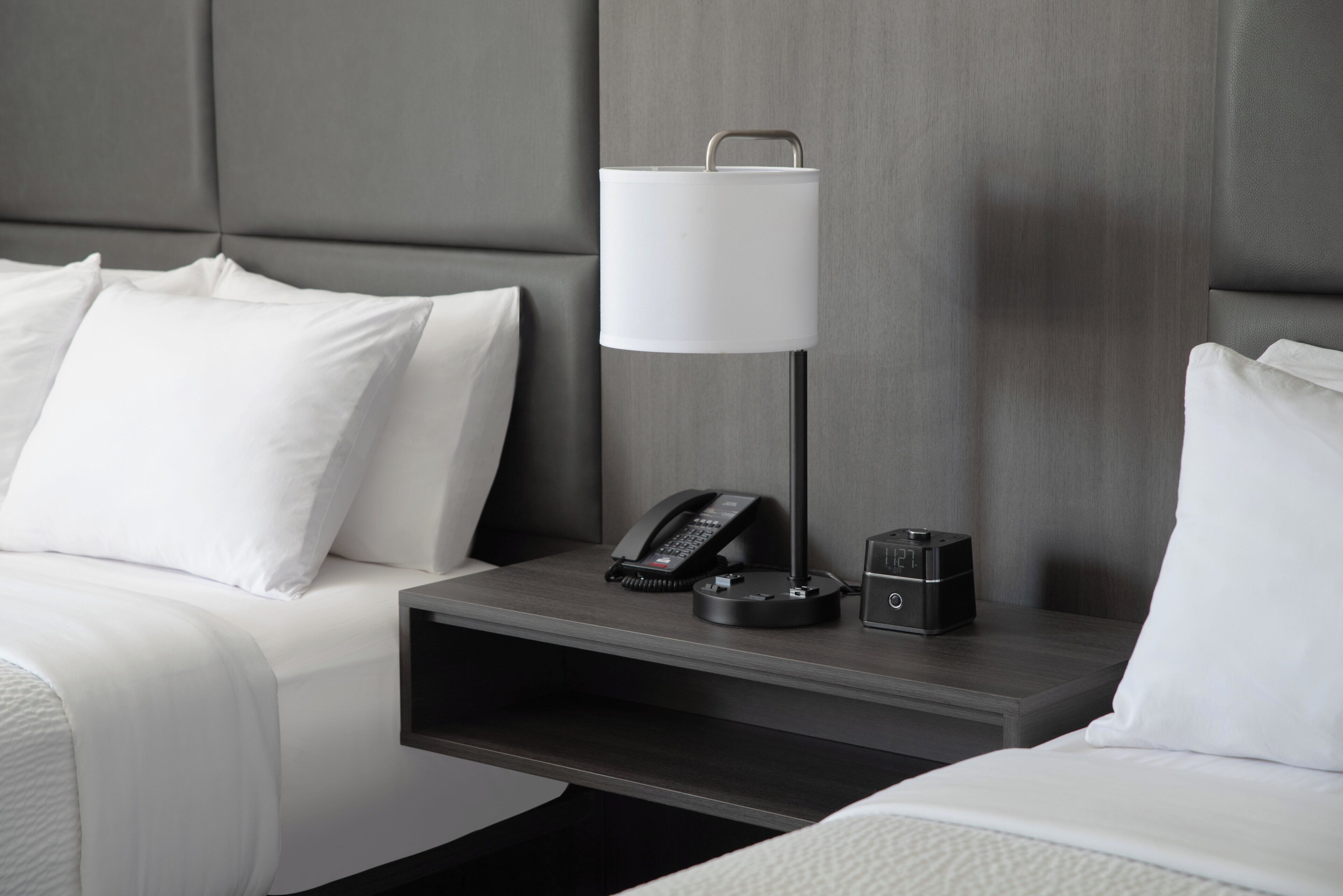 Una habitación de hotel con una cama, una mesita de noche con lámpara, un teléfono y un altavoz pequeño.
