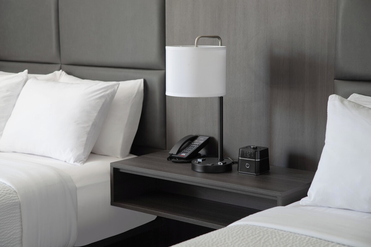 A hotel room with a bed, a bedside table with a lamp, a phone, and a small speaker.