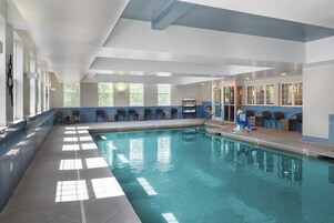 Una piscina interior con agua cristalina, rodeada de sillas y una zona iluminada por el sol con ventanas.