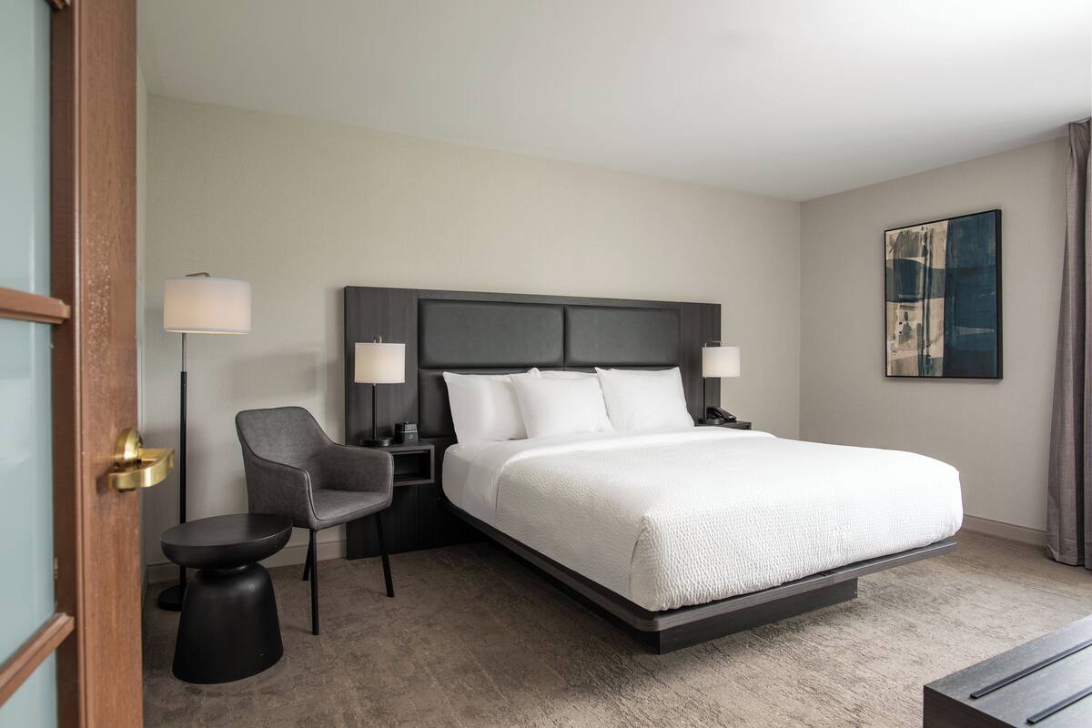A modern hotel room with a large bed, a desk, a chair, and a lamp.