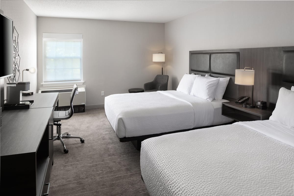 A hotel room with two beds, a desk, a chair, a lamp, and a TV.