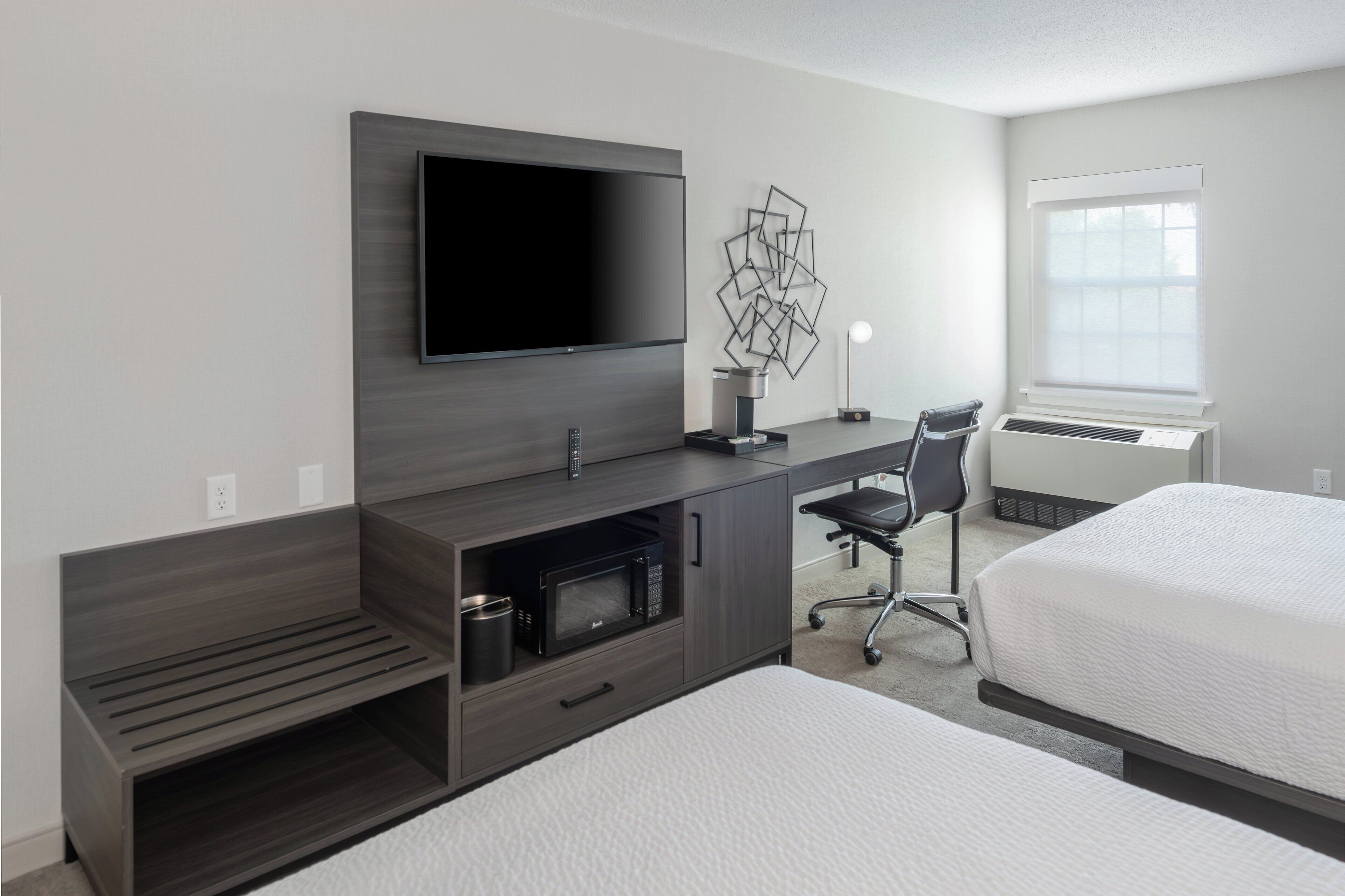 Habitación de hotel con televisor de pantalla plana montado en la pared, escritorio con silla, microondas y cama con colcha blanca.
