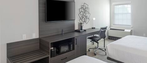 Habitación de hotel con televisor de pantalla plana montado en la pared, escritorio con silla, microondas y cama con colcha blanca.