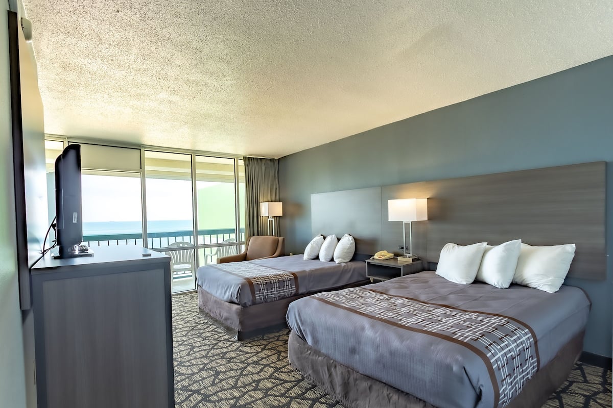 Quarto de hotel com duas camas, uma escrivaninha, um telefone e vista para o oceano.
