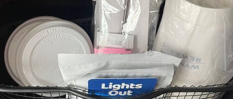 Una cesta con artículos de aseo, incluyendo una taza, una botella de crema vegetal, una caja de pañuelos de papel y una caja con la etiqueta 'Lights Out'.