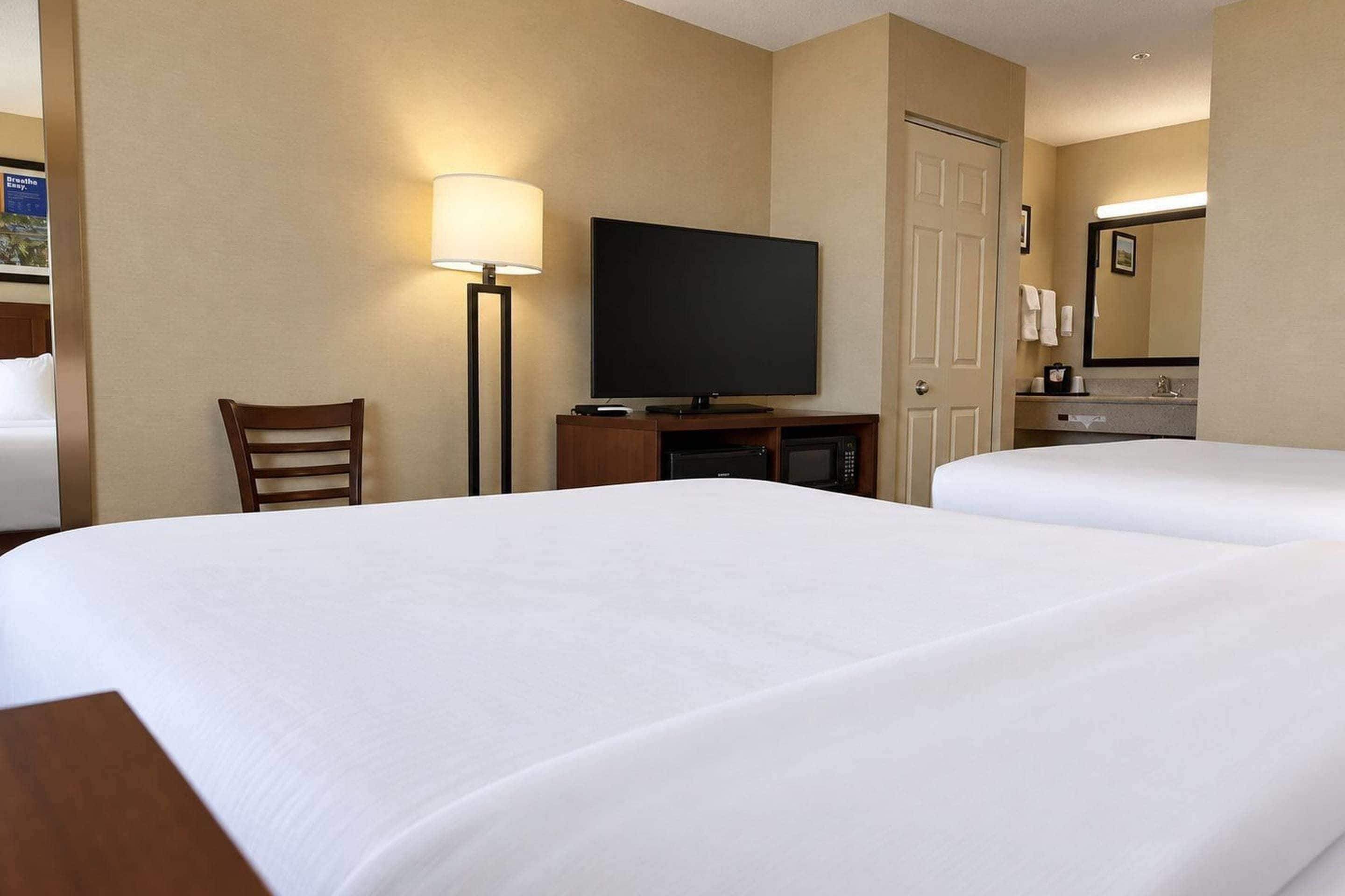 A hotel room with two beds, a television, a desk with a lamp, a chair, and a bathroom with a mirror.