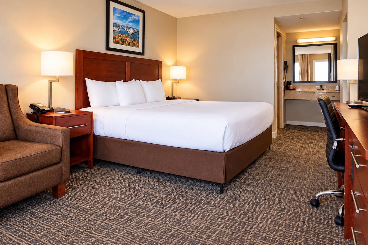 A hotel room with a large bed, two bedside tables with lamps, a chair, a desk with a chair, and a mirror.