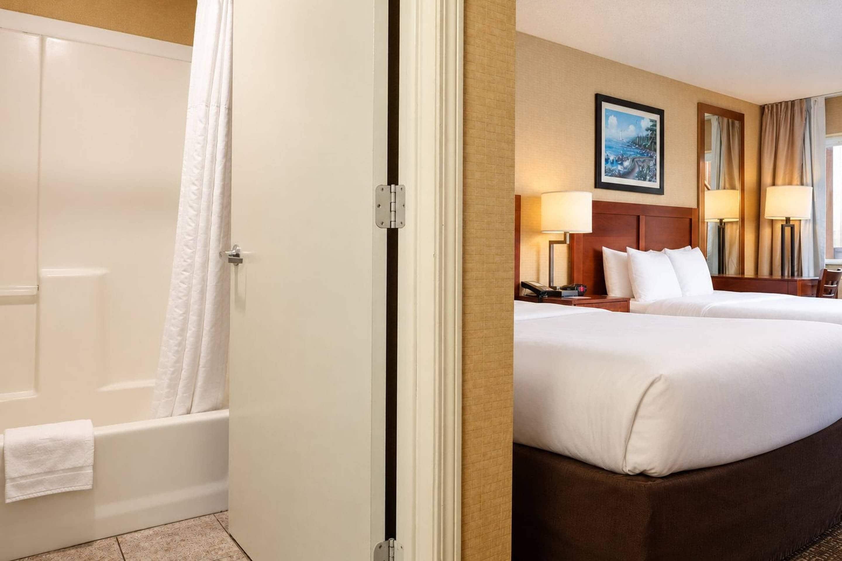 Ein Hotelzimmer mit einer Badewanne, zwei Betten, einem Kopfteil, Lampen, einem Gemälde und einem Fenster mit Vorhängen.