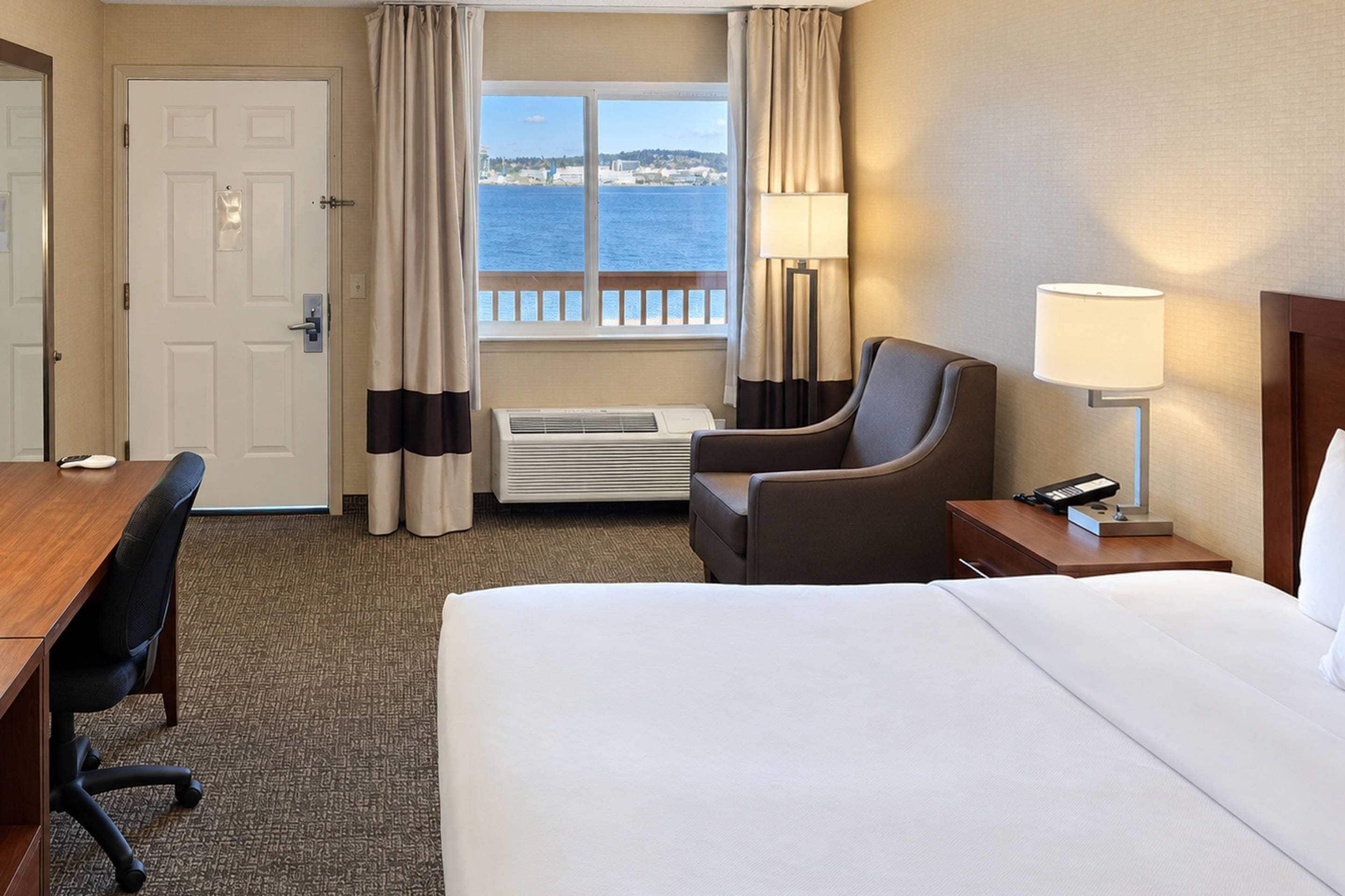 A hotel room with a bed, a desk, a chair, a nightstand, a lamp, and a window with a view of the sea.