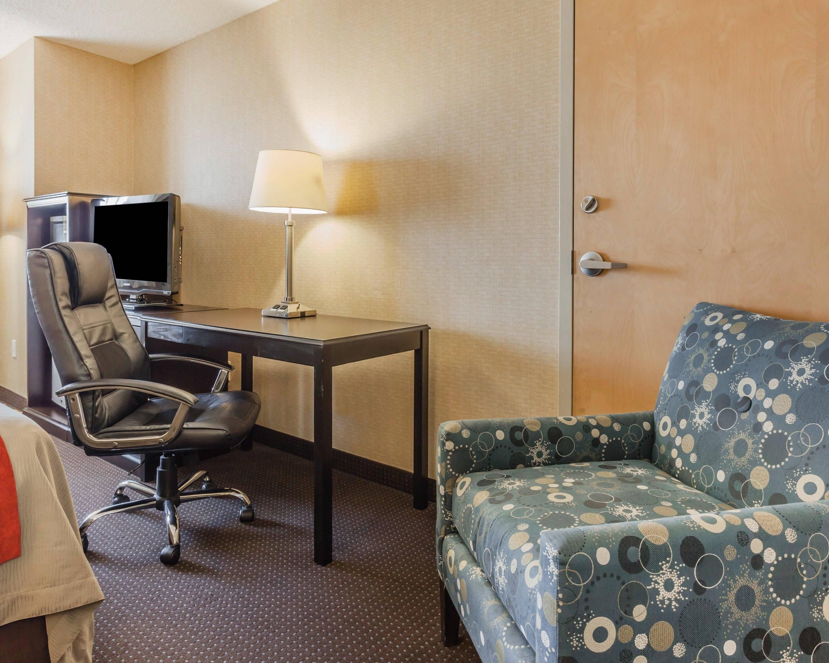 A hotel room with a desk, chair, TV, lamp, and a patterned sofa.