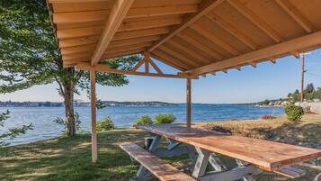 Une table de pique-nique en bois, placée sous un abri au bord d’un lac, offre une vue dégagée sur l’eau et les bâtiments situés au loin.