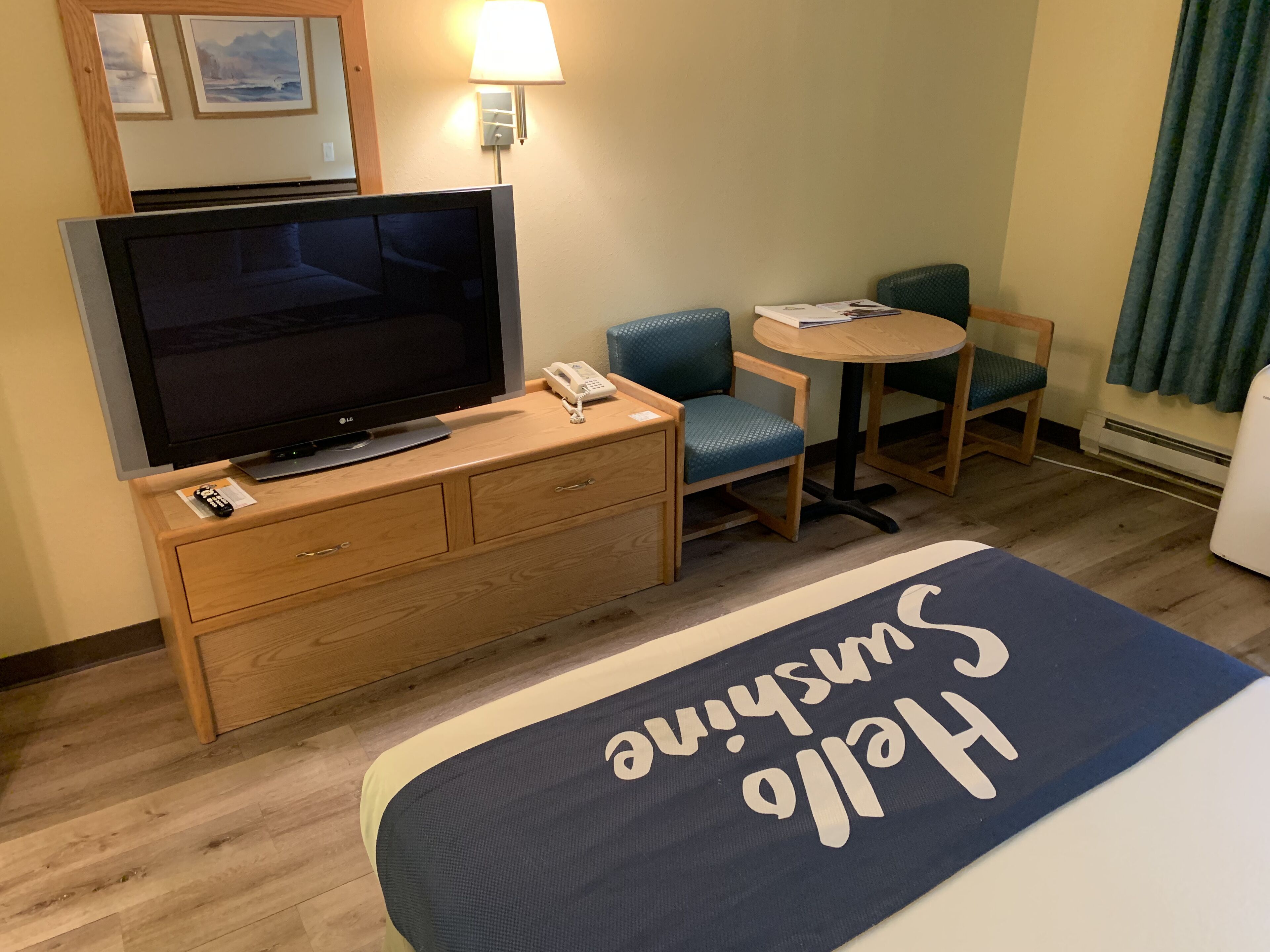 A hotel room with a bed, a TV on a wooden dresser, a desk with a chair, and a lamp.