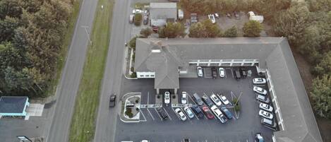 An aerial view of a parking lot with multiple rows of parked cars, a building with a sloped roof, and a road adjacent to the parking area.