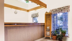 A reception area with a wooden reception desk, a clock, a window with a floral curtain, and potted plants. - Days Inn by Wyndham Ocean Shores (Ocean Shores)