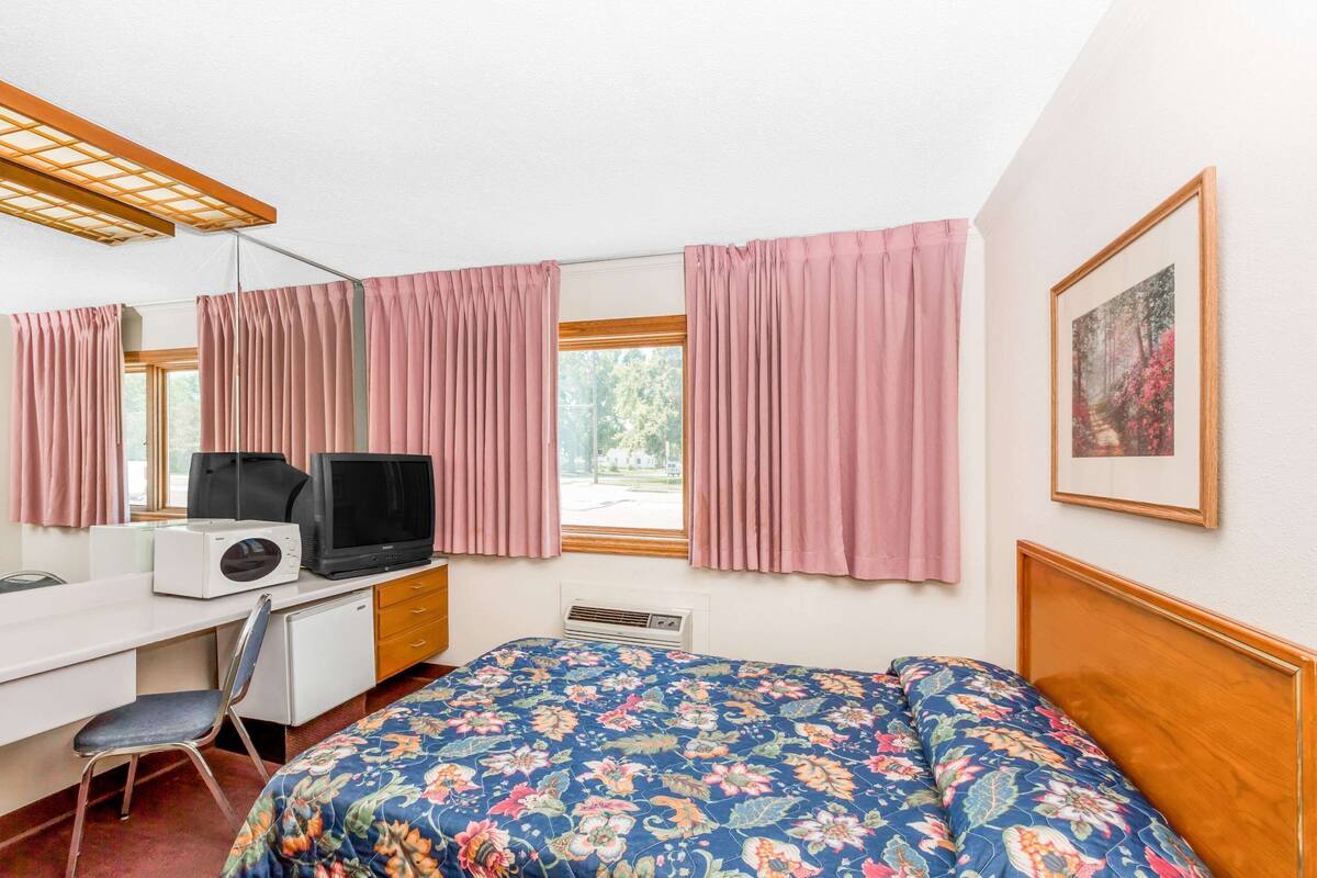 A room with a bed, a desk with a television, a chair, and a window with curtains.