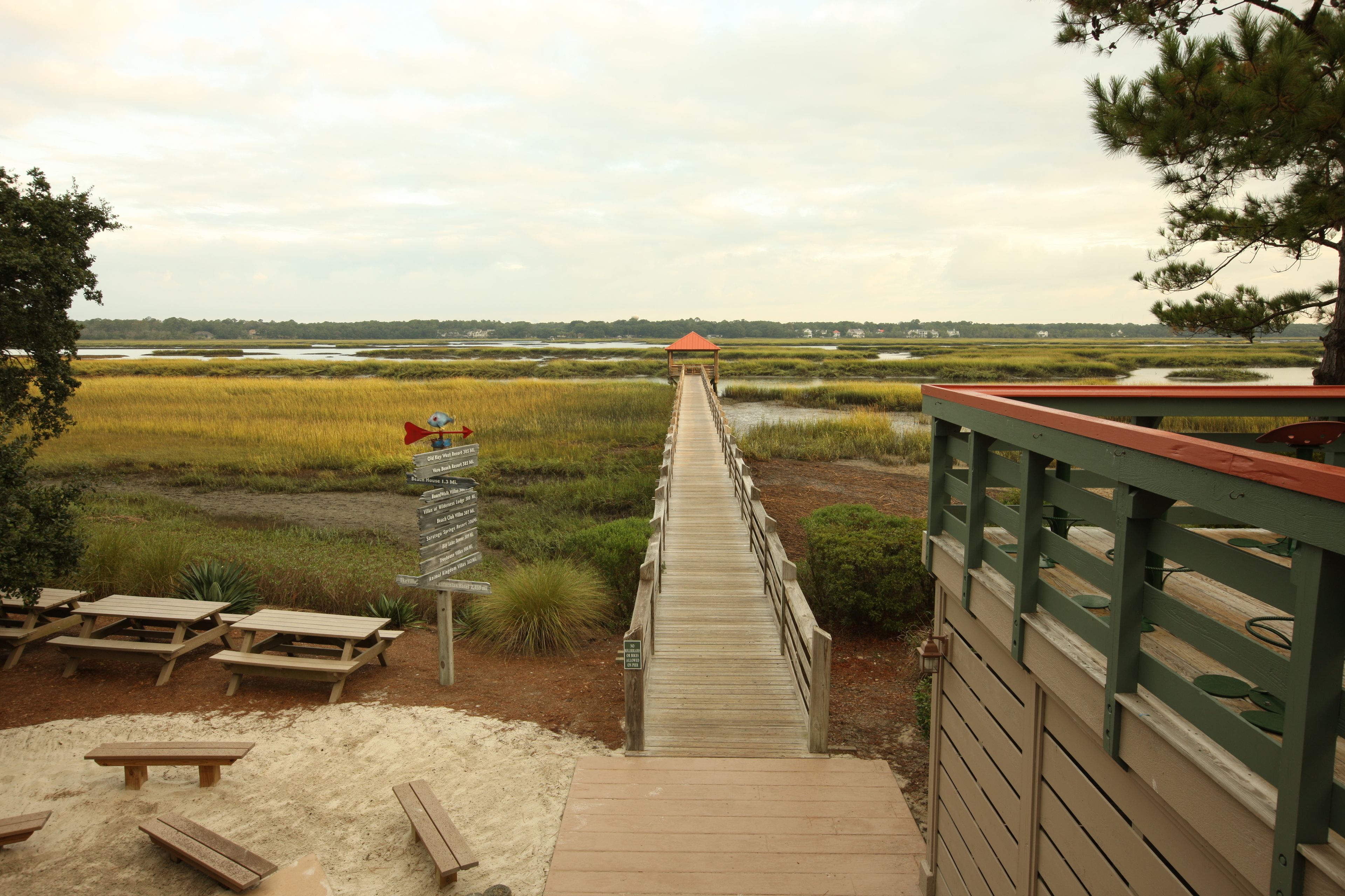 A wooden boardwalk leading to a gazebo, surrounded by sand and vegetation.