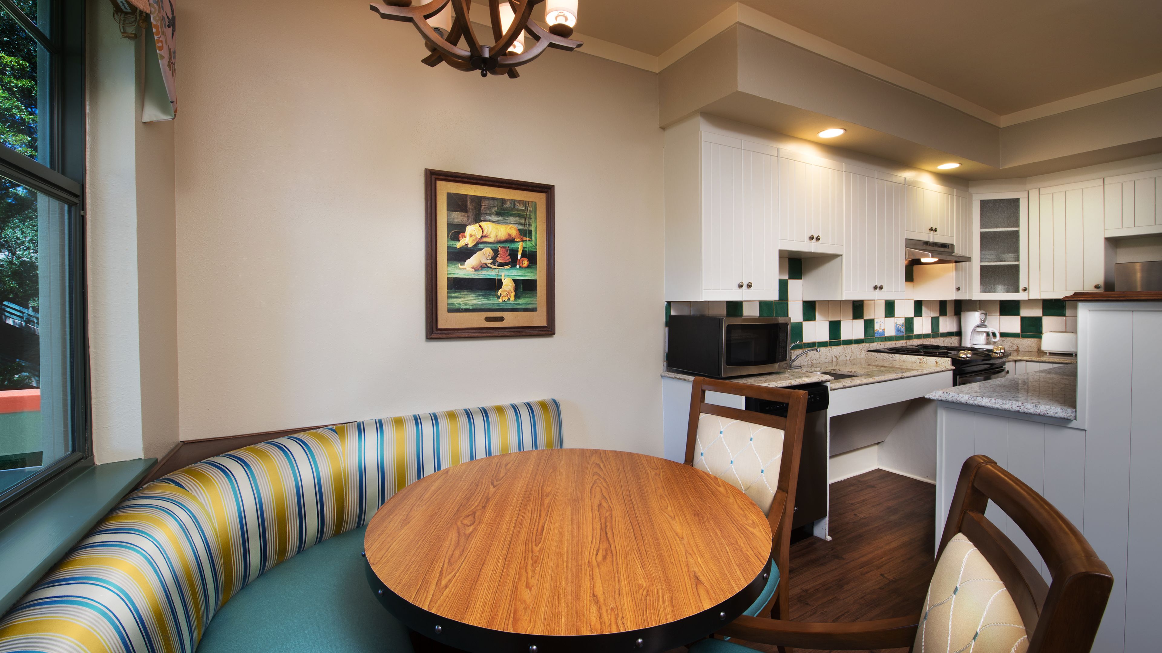 A compact kitchen with white cabinets, a microwave, and a stove. There's a wooden dining table with chairs and a striped bench. A framed picture hangs on the wall.
