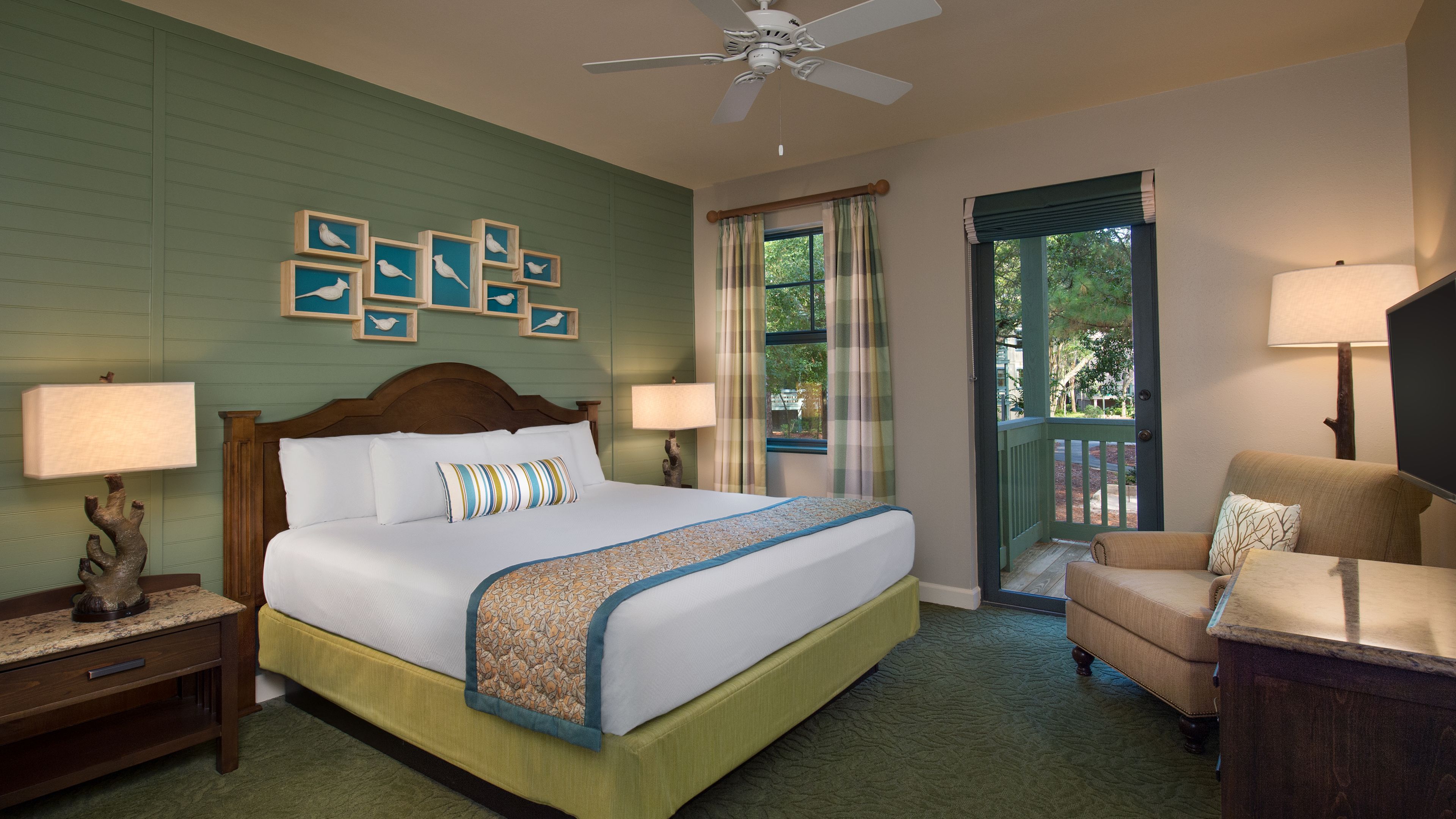 A hotel room with a large bed, two bedside tables, a chair, a ceiling fan, and a view of the outdoors through a window.