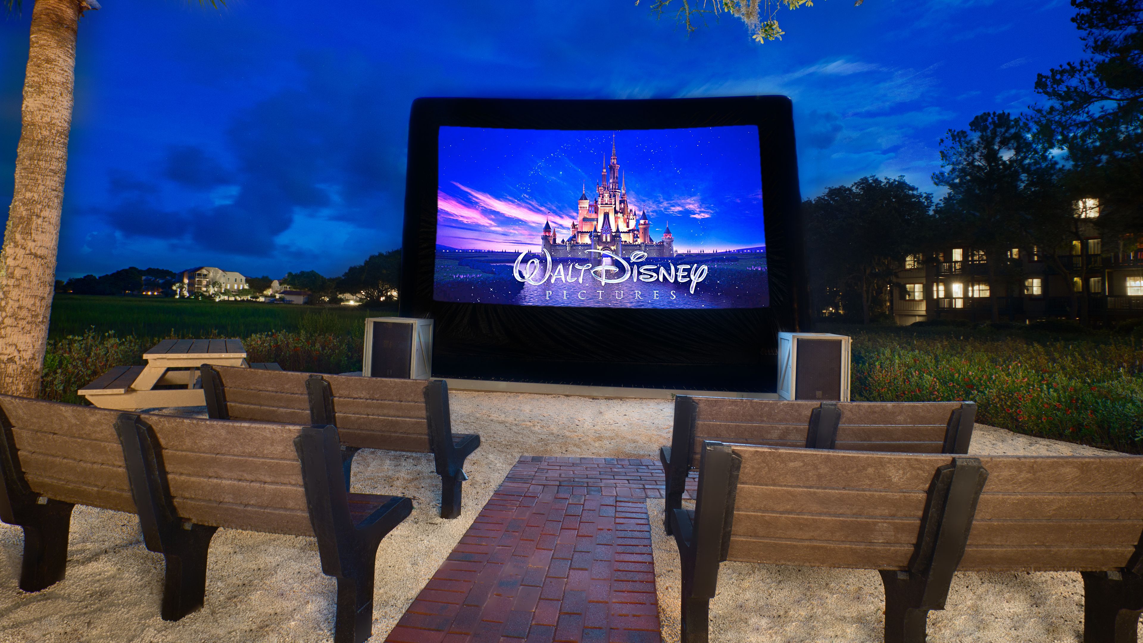 Una pantalla de cine al aire libre con la imagen de un castillo de Disney, bancos y un camino de ladrillo.