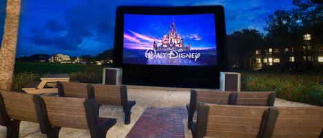 Una pantalla de cine al aire libre con la imagen de un castillo de Disney, bancos y un camino de ladrillo.