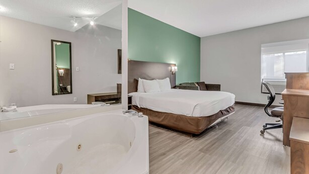 A modern hotel room with a large white bathtub, a bed with white bedding, a desk with a chair, and a mirror on the wall. - Baymont by Wyndham N Myrtle Beach/Park & Sports Complex Area (North Myrtle Beach)