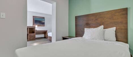 A hotel room with a large bed, a wooden headboard, a desk with a chair, and a view through an open doorway.