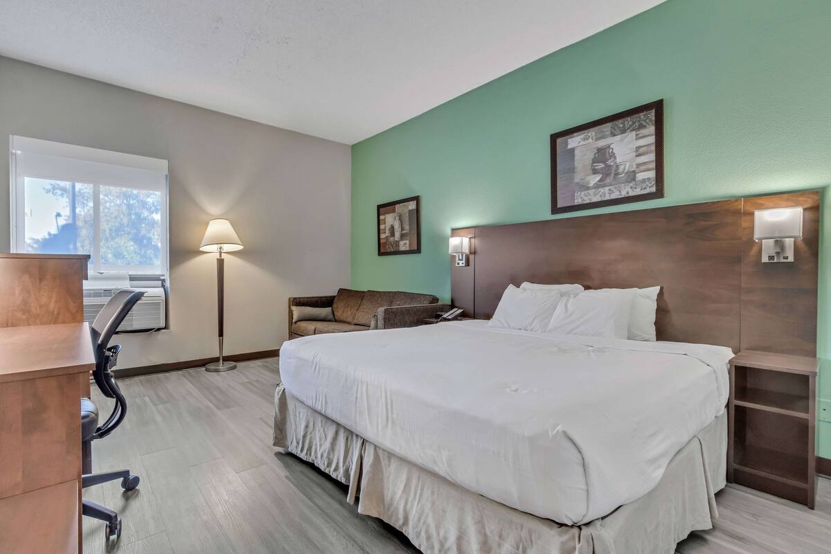 Une chambre d’hôtel avec un grand lit, un bureau, une chaise, une lampe sur pied et une fenêtre équipée d’un appareil de climatisation.