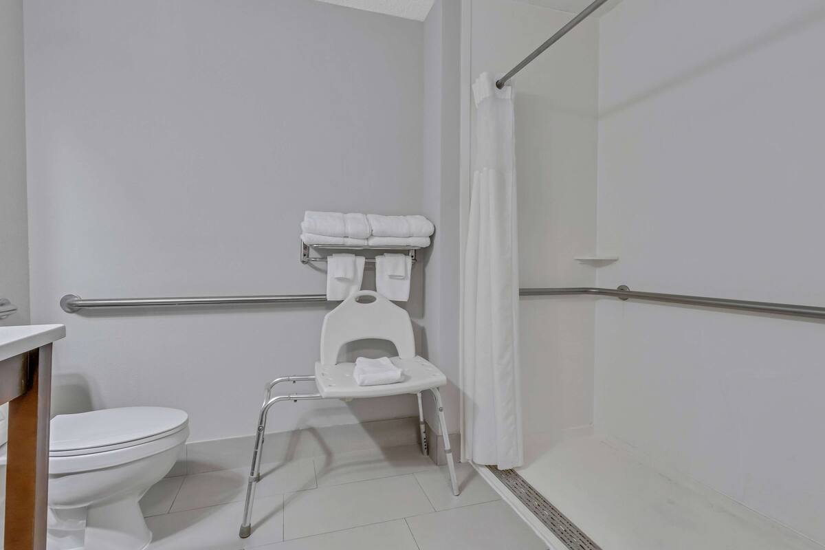 Une salle de bain avec des toilettes blanches, une douche blanche dotée d’une porte en verre et une chaise blanche avec une serviette.