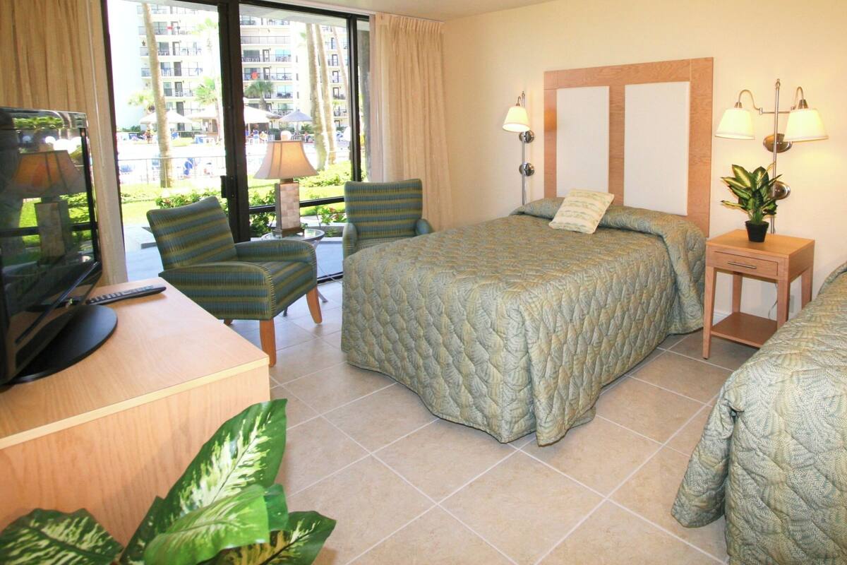 A hotel room with two beds, a desk with a TV, a chair, and a small table with a plant.