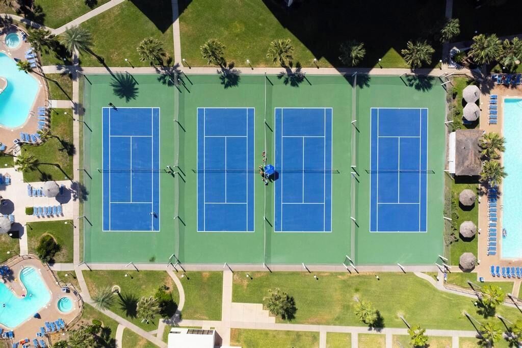 An aerial view of a tennis court complex with multiple courts, a swimming pool, and surrounding greenery.