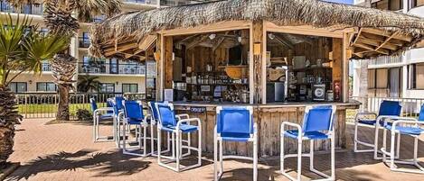 Zona de bar con techo de paja, rodeada de sillas azules y un ambiente junto a la piscina.