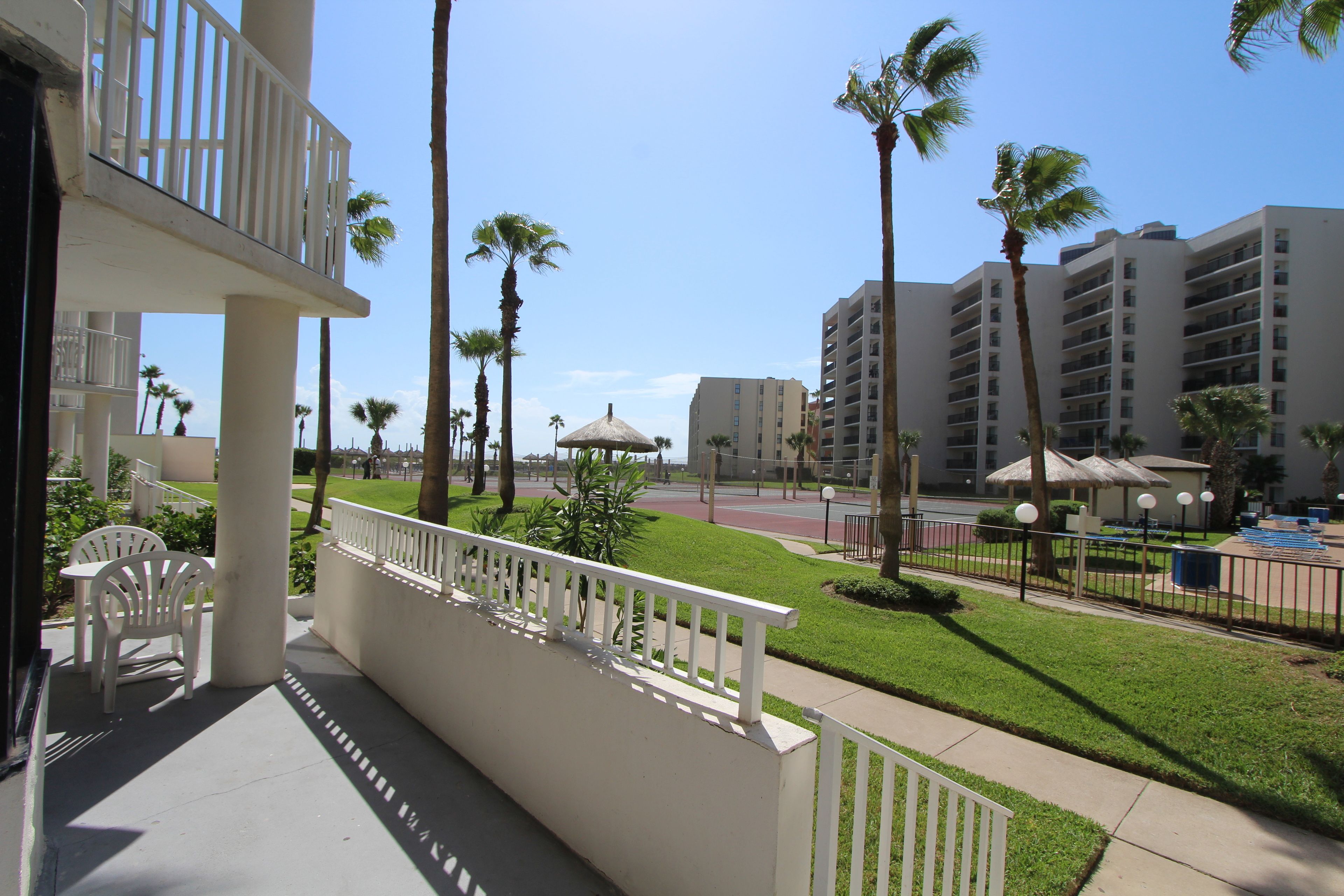 A hotel balcony with white railings, a grassy lawn, palm trees, and a view of a multi-story building.