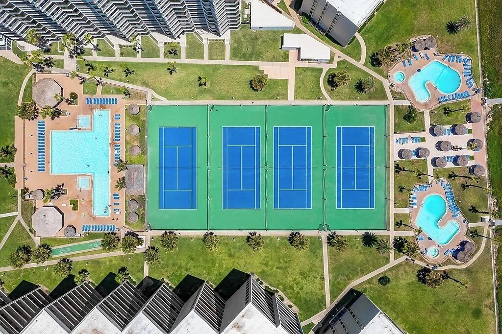 An aerial view of a resort with a tennis court, swimming pools, and a landscaped area with lounge chairs.