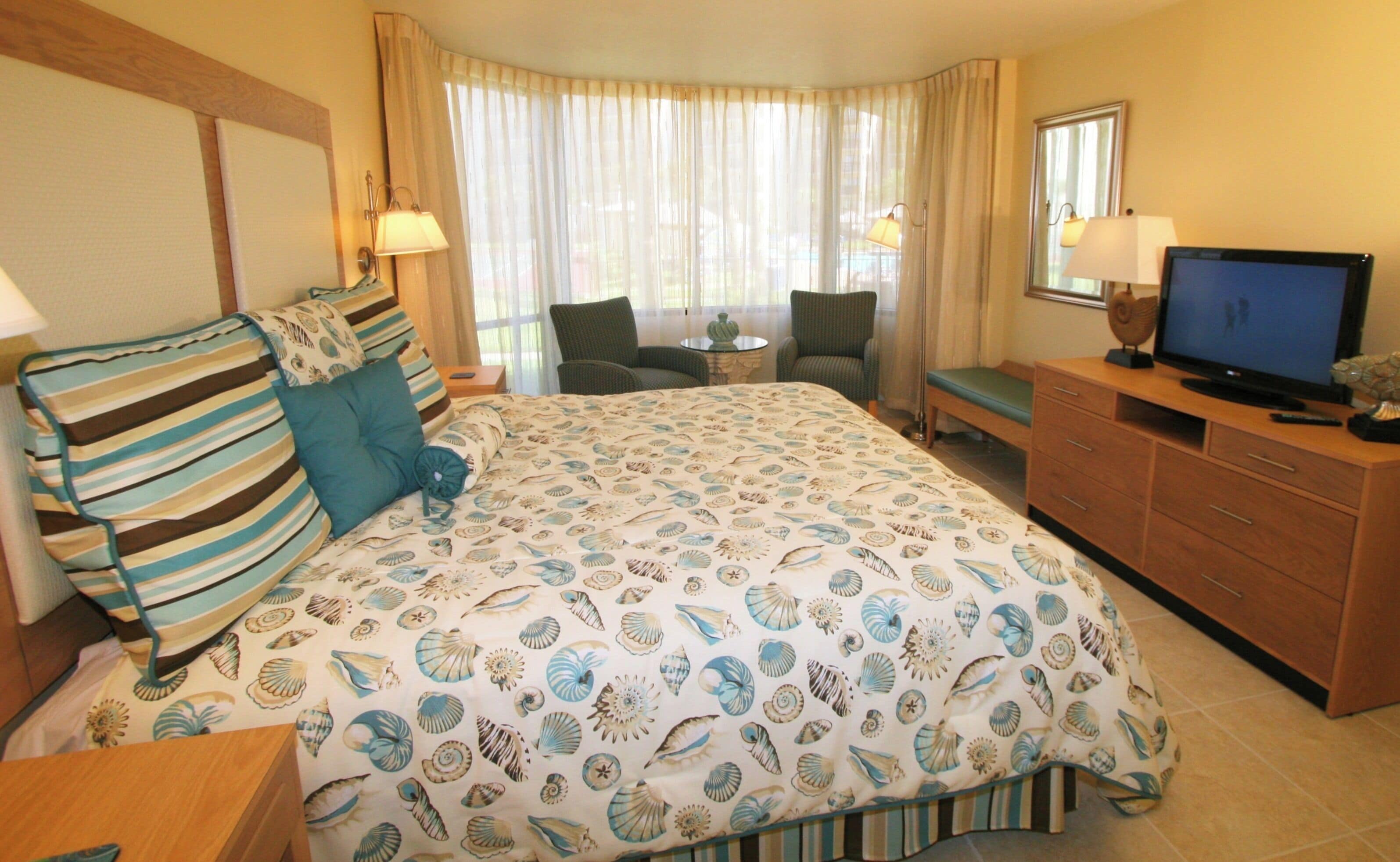 A hotel room with a large bed, a desk with a TV, a chair, and a small table.