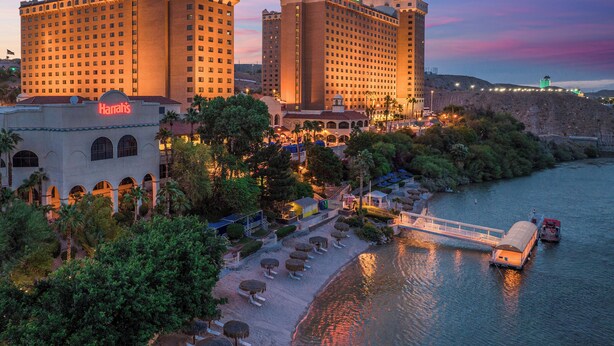 A large hotel complex with multiple buildings, a riverside promenade with sun loungers, and a boat dock.