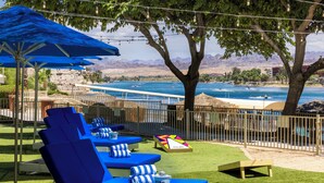 A poolside area with blue lounge chairs and umbrellas, a wooden deck, and a view of a body of water with mountains in the background.