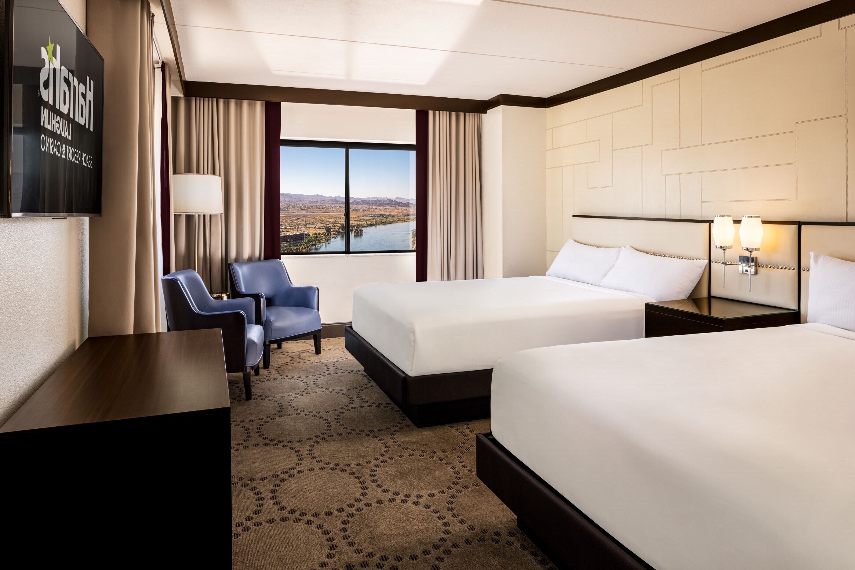 A hotel room with two beds, a desk, a chair, a TV, and a view of a lake.