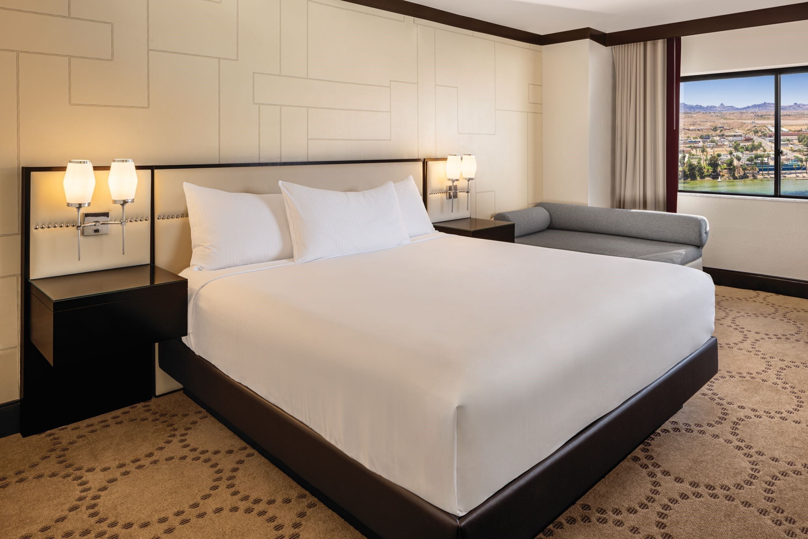 Een hotelkamer met een groot bed, twee bedlampjes, een bank en uitzicht op een landschap door een raam.