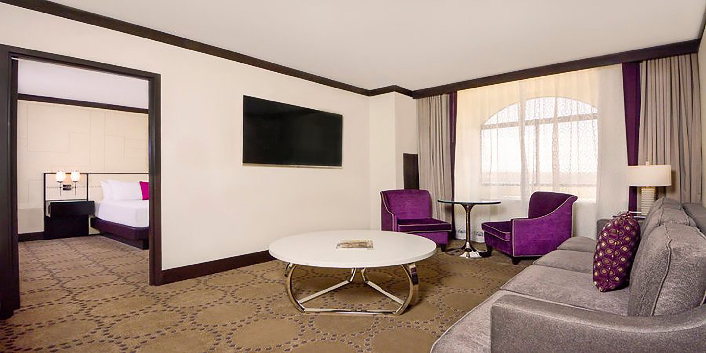 A hotel room with a bed, a round table, two purple armchairs, a grey sofa, and a TV mounted on the wall.