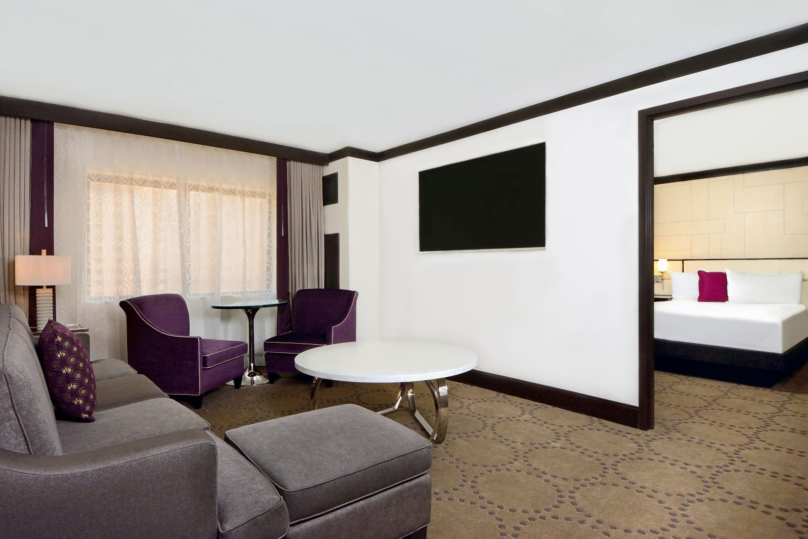 Quarto de hotel com sofá cinza, poltrona roxa, mesa redonda pequena e cama com cabeceira branca.