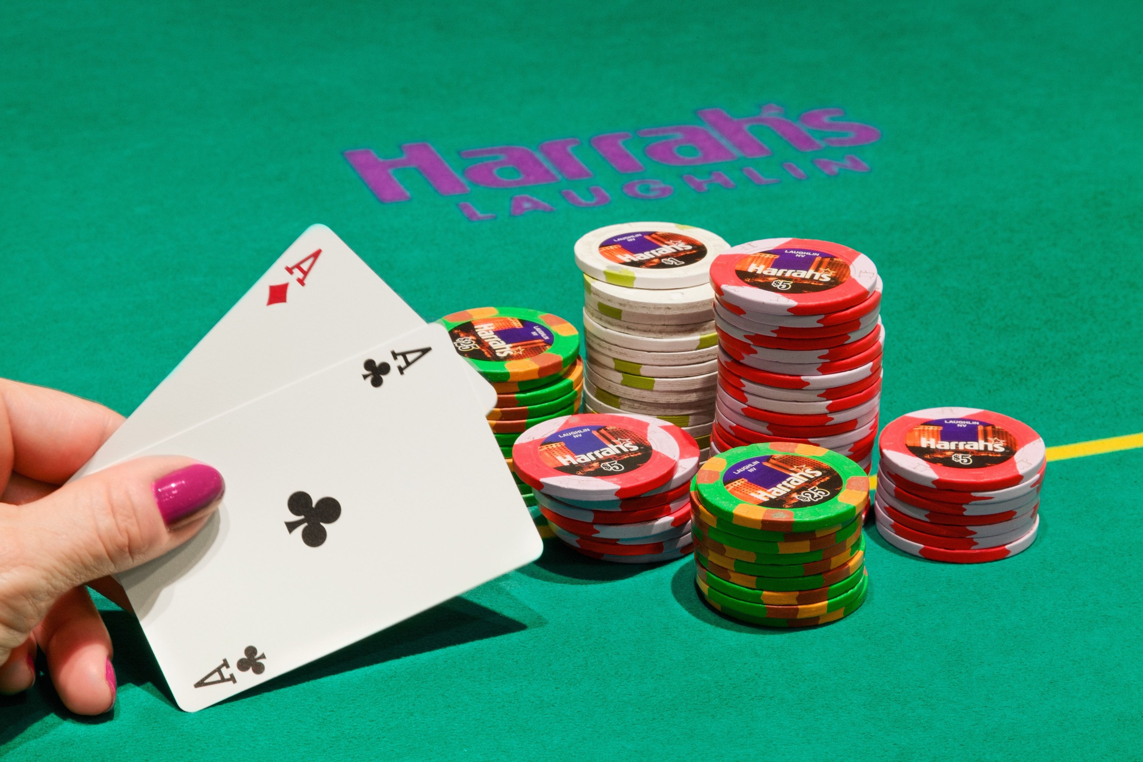 Une main tenant une carte à jouer ornée d’un trèfle, des piles de jetons de poker et une table de jeu recouverte de feutre vert sur laquelle on peut lire le texte « Harrah’s Laughlin ».