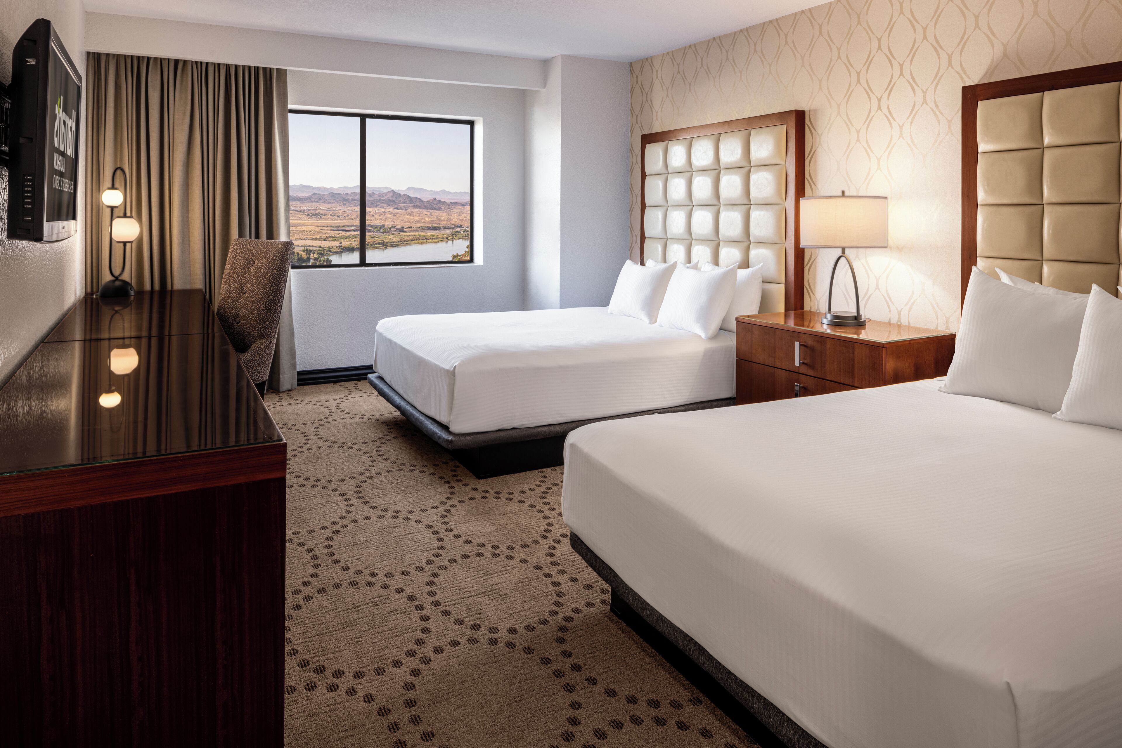A hotel room with two beds, a desk, a TV, and a window with a view of the mountains.