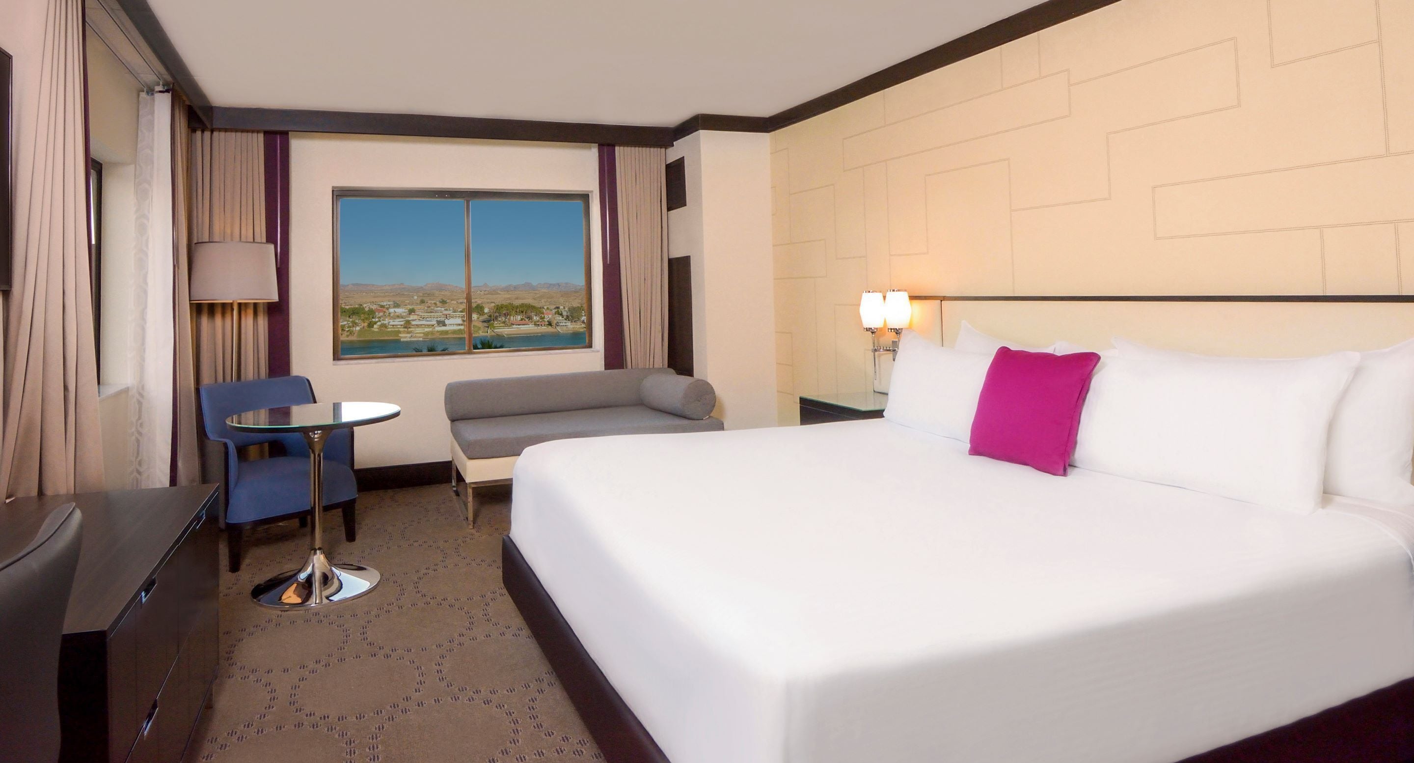 A hotel room with a large bed, a small table, a sofa, and a view of a landscape through a window.