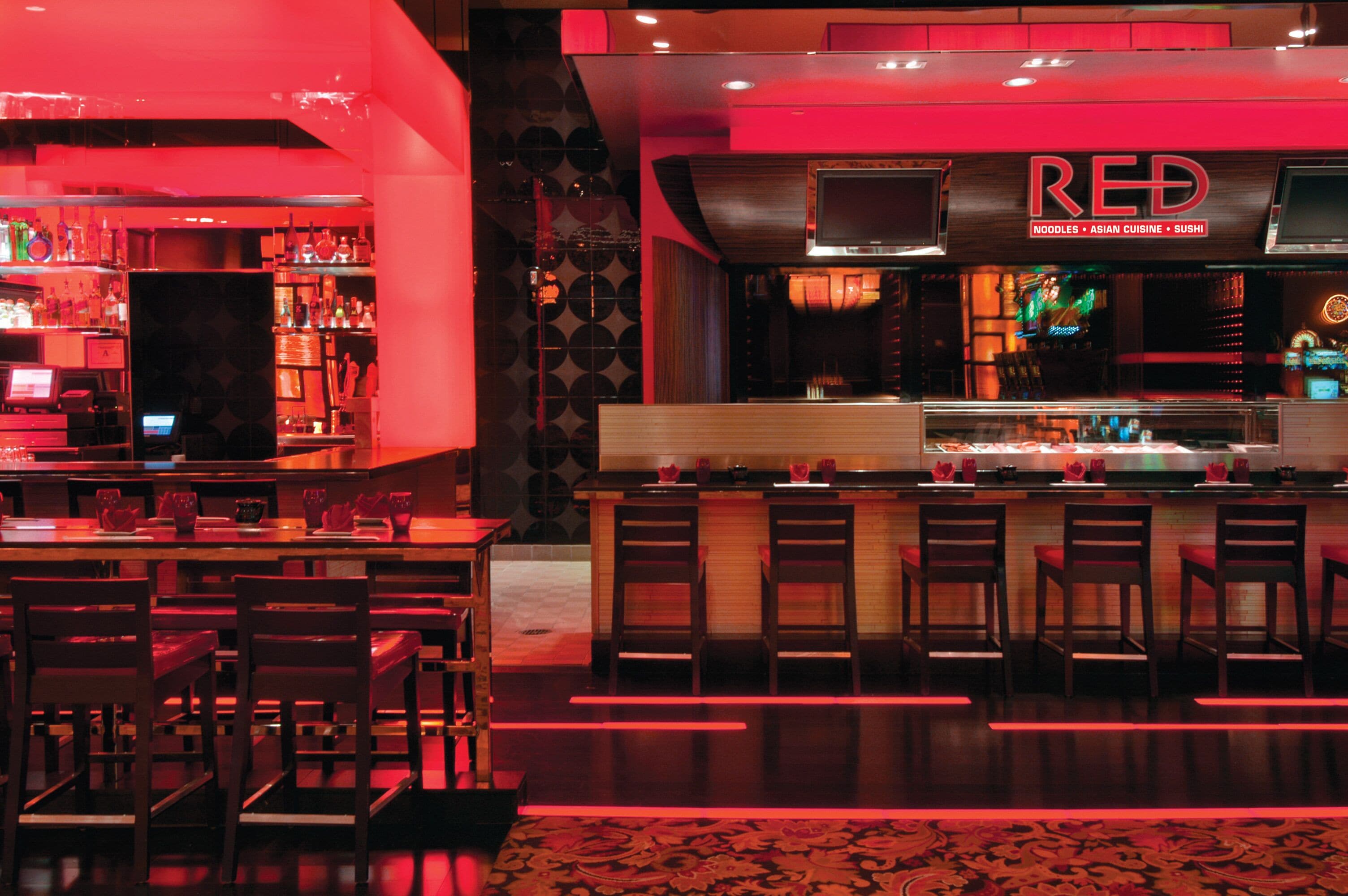 L’intérieur d’un restaurant comprend un bar, plusieurs espaces aménagés pour accueillir les clients et une enseigne bien visible, de couleur rouge, qui indique le type de cuisine proposée.