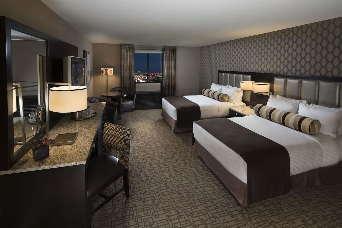 Quarto de hotel com duas camas, uma escrivaninha com abajur, uma cadeira, um espelho e vista para a cidade à noite.