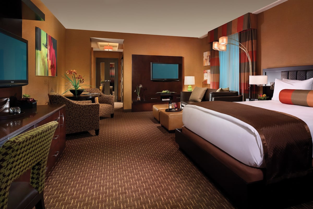 Quarto de hotel com uma cama grande, uma mesa com televisão de tela plana, área de estar com cadeiras estampadas e piso coberto por carpete.
